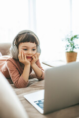 A little girl, 5-6 years old, reclines on a sofa, wearing headphones and watching a laptop screen. Concept: technology-infused relaxation, online education, computer entertainment - obrazy, fototapety, plakaty