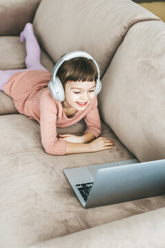 A laughing little girl, aged 5-6 years old, lying on a sofa as she watching a laptop. Concept: technology-infused relaxation, online education, computer entertainment