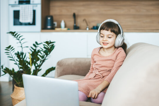 A smiling 5-6 years old girl watching a laptop screen, sitting on a beige couch. Concept: technology-infused relaxation, online education, computer entertainment