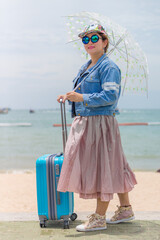Smiling female tourist holding umbrella and pulling modern blue rolling hard baggage. She was walking on the sidewalk while visiting the sea beach.