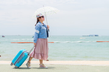 Female tourist holding umbrella and pulling modern blue rolling hard baggage. She was walking on the sidewalk while visiting the sea beach.