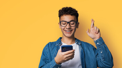 Smiling funny young man wear braces eye glasses hold typing text look read cell phone cellular smartphone cellphone, crossing fingers, praying hoping for good luck, isolated orange yellow background.