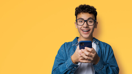 Excited surprised shocked astonished happy curly haired funny young man wear braces glasses spectacles open mouth hold typing cell phone cellular smartphone cellphone isolated yellow background