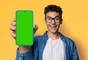 Shocked happy man wear glasses braces, open mouth, show cell phone, smartphone, mobile phone, cellphone green chroma key mockup mock screen, isolated yellow wall background. Online offer ad.