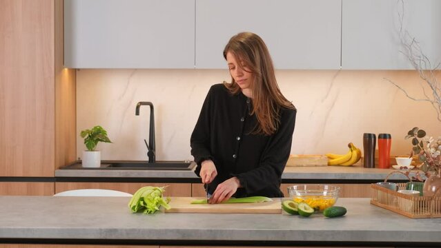 Young woman standing at kitchen and slicing celery for healthy salad.