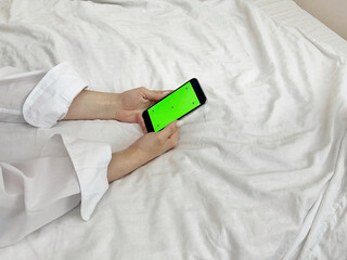 Female hands in a white shirt against the background of a bed hold a phone with a green screen. Mobile Phone Day. Mobile Phone Give Up Day