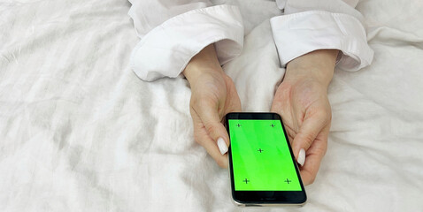Female hands in a white shirt against the background of a bed hold a phone with a green screen....