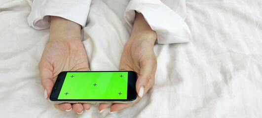 Female hands in a white shirt against the background of a bed hold a phone with a green screen. Mobile Phone Day. Mobile Phone Give Up Day