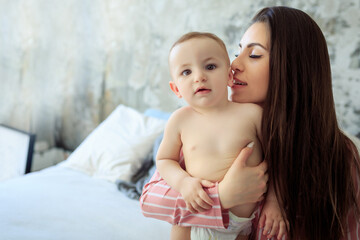 Young Caucasian brunette mother embracing, kisses her baby boy son sitting on white bed in sunny...