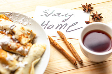 Tasty pancake rolls, tea cup and card with message Stay Home on wooden table. Top view. Quarantine concept
