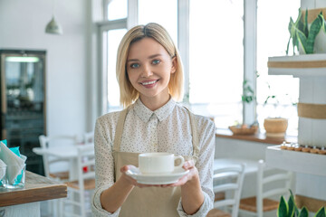 Smiling woman serving coffee inside a coffee shop. Happy coffee shop owner holding a cup of coffee inside the shop. Serving decaf sugar free non-lactose milk hot beverage cacao chocolate in cafe
