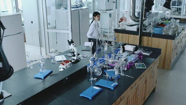 Full high angle shot of Asian female employee of research chemical laboratory working on new synthetic compounds - typing on laptop, then walking over to flasks with liquids and adding reagent