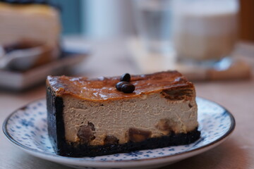 A piece of espresso macadamia cheese cake topped with coffee beans on the plate with blur of blueberry crepe cake and dirty latte coffee.