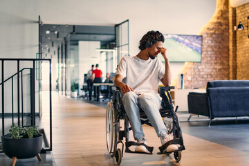 A African-American teenager in a wheelchair sits sadly amidst the bustling backdrop of a modern startup office, surrounded by his business colleagues.
