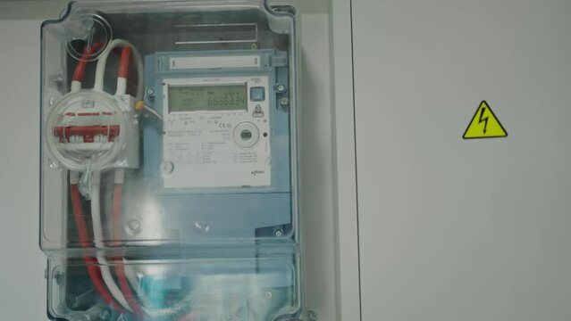 Clear shot of an advanced electricity meter housed in a transparent box next to a warning sign. Modern Electric Meter in Transparent Box On Office Wall