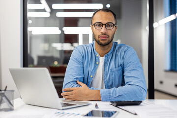 Portrait of young hispanic man in glasses and shirt, office worker and student sitting at desk with...