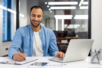 Smiling young hispanic male student sitting in office, working and studying remotely via laptop, making notes in notebook