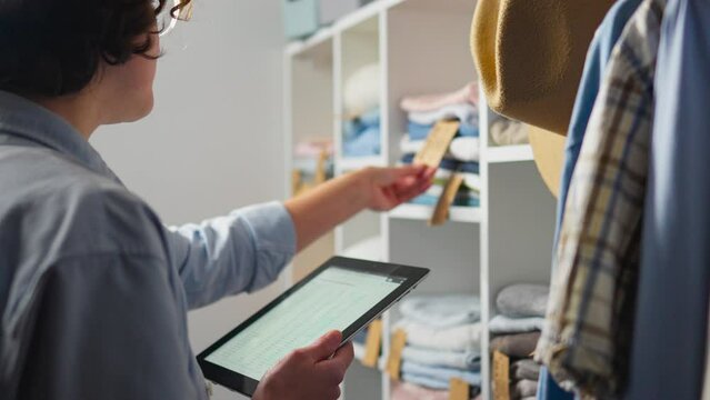 A young warehouse worker keeps track of clothing items using a modern touchscreen tablet, entering digital data into spreadsheets. The employee will use modern technologies in accounting