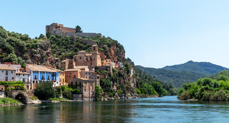 Fototapeta na wymiar Idyllic riverside village with terracotta-roofed houses nestled on a lush hillside, crowned by a majestic fortress, under the azure sky.