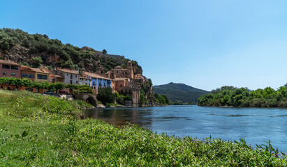 a serene river flowing past a hilltop village, with the architecture of the village merging seamlessly into the lush, natural landscape under a clear blue sky