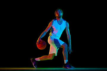 Muscular, athletic young man, basketball player in motion with ball, practicing against black background in neon light. Concept of sport, competition, active and healthy lifestyle, game