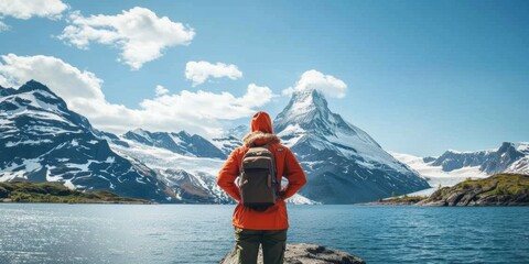 A lone adventurer in a vibrant orange jacket stands gazing at majestic snow-capped mountains reflected in the tranquil lake waters under a clear blue sky. - obrazy, fototapety, plakaty