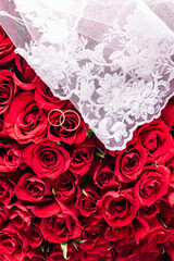 Background image for wedding design or mockup. Two gold wedding rings on a red roses bouquet background. white bridal veil. Top view. A copy space.