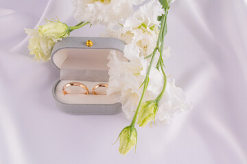 Two gold wedding classic rings in a gray velvet box on a white satin fabric with fresh white flowers. Wedding concept. Romantic design.