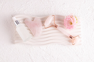 A white matte bottle of cosmetics, a rose quartz massage roller and a gua sha scraper lie on an embossed podium. Top view.