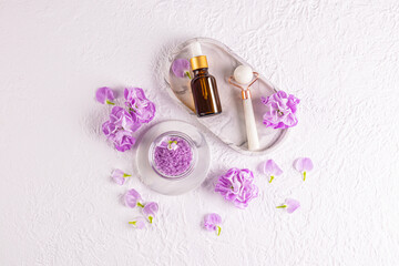 A set of cosmetic oil or serum for massage, a roller made of white stone on a tray. Top view. lilac delicate flowers and bath salts. Spa treatments.