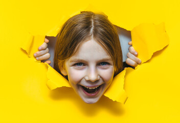 A funny laughing red-haired girl looks out through a hole in yellow paper. The concept of surprise, smile, joyful mood from what he saw. Discounts, sales, surprise. Copy space.