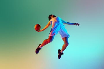 Fototapeta na wymiar Bottom view dynamic image of young man, basketball player in motion, throwing ball against gradient background in neon light. Concept of sport, competition, active and healthy lifestyle, game