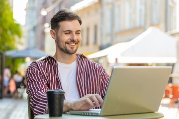 Brunette handsome man working study education online distant job with laptop in city sunny street cafe restaurant browsing website chatting outdoors during break. Smiling guy tourist drinking coffee