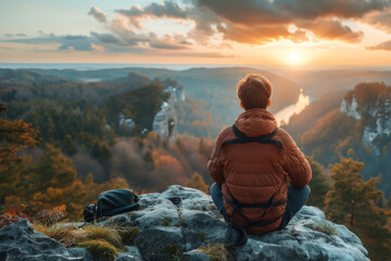 A young Caucasian man  sitting on a rocky cliff overlooking a scenic mountain landscape at sunset - Powered by Adobe