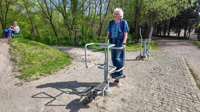 Old gray-headed male senior in his seventies (75) in blue  jeans pants and jacket exercising on a hip twister in the outdoor fitness gym in a public park and his wife resting on a bench. 4K video
