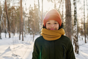 Adorable boy in knitted yellow scarf and brown beanie hat looking at camera with smile while standing in natural environment