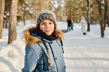 Cute smiling boy in warm blue winter jacket and knitted beanie hat looking at camera while standing...
