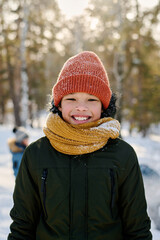 Cute cheerful African American boy in winterwear looking at camera with toothy smile while standing in natural environment on winter day