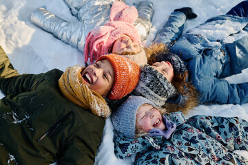 Several intercultural kids in beanie hats and warm jackets lying on snow and laughing while...