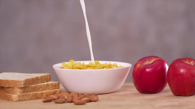 Pouring milk into  a bowl of cornflakes, red apple and whole wheat breads on the table