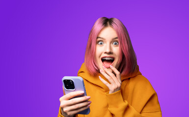 Shocked astonished very happy pink woman wear braces, orange hoodie open mouth, using hold hand cell phone, smartphone, mobile phone, isolated violet purple background. Online offer, dental care ad.