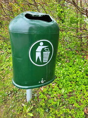 Grüner Müllbehälter im Freien mit einem weißen Symbol. Der Mülleimer steht auf Gras und ist von verschiedenen grünen Pflanzen umgeben, was auf eine parkähnliche oder natürliche Umgebung hindeutet.