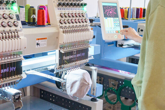 Man's hand pointing onthe screen of embroidery 15 Needle Compact Embroidery Machine next to colorful threads  and cap. Setting up a modern embroidery machine. Turkey, Istanbul, April 20, 2024.