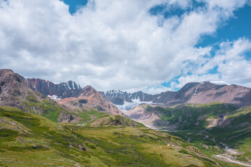 Colorful vast scenery of wide green alpine valley with panoramic view to large snow-capped top, sharp rockies, rocky pointy peak, snowy mountain range and big glacier tongue far away under cloudy sky. - 793991652