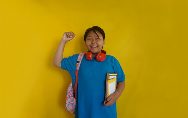 Asian little girl with headphones on neck, holding book, clenching fist and smiling looking at...