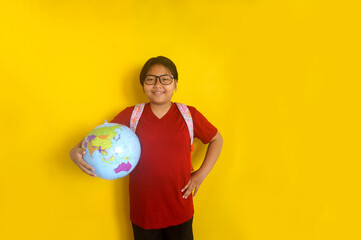Asian little girl student wear glasses and backpack holding globe in one hand other on waist isolated over yellow background