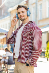 Happy relaxed Caucasian bearded adult man dancing in wireless headphones choosing, listening energetic disco rock n roll music outdoors. Young guy standing on urban city sunshine street. Vertical