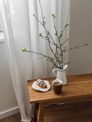 Cozy aesthetic coffee break - a cup of coffee, a croissant, a jug with spring branches on a wooden...