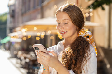 Happy smiling teenager girl using smartphone typing text messages browsing internet social media web app working chatting online outdoor. Redhead child tourist walking in urban city street. Lifestyles