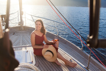 Beautiful woman traveling and drinking wine on yacht at sea. Summer vacation at sunset. Happy girl in straw hat relaxing, enjoying holidays with wineglass of alcohol-free beverages. Lifestyle moment - obrazy, fototapety, plakaty